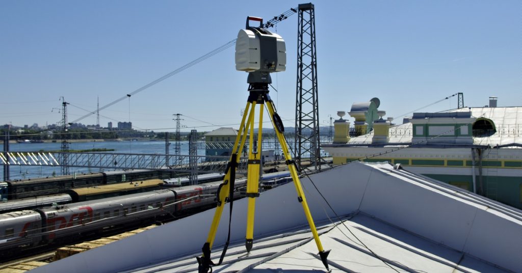 A laser scanning device sitting on top of the roof of an industrial building. There are lines of trains in the background.