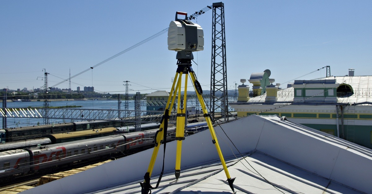 A laser scanning device sitting on top of the roof of an industrial building. There are lines of trains in the background.