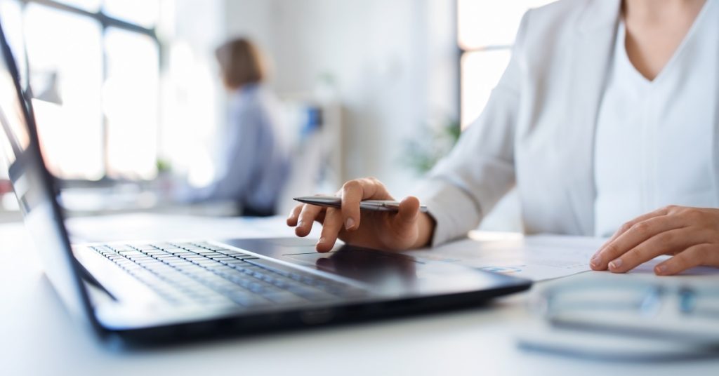 A woman with a pen in her hand using a laptop touchpad. She's in a business environment with someone working behind her.