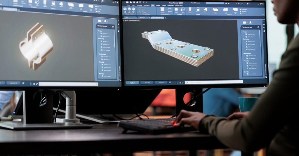 A woman sitting at two monitors, each with 3D components on them. She's currently working on both of them.