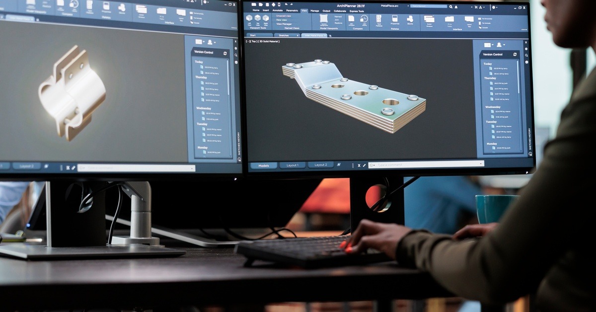 A woman sitting at two monitors, each with 3D components on them. She's currently working on both of them.