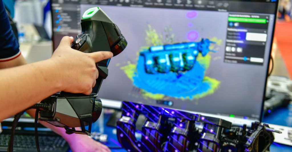 A person using a 3D scanner on an engine part. There are purple scan lines on the object and a screen showcasing it.