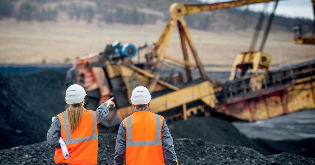 Two workers in bright vests and hard hats looking at some mining equipment in the distance. One of them is pointing at it.