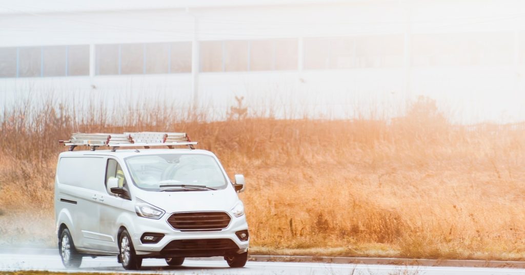 A service van with a roof rack driving down a wet road. There is a large warehouse building behind the van.