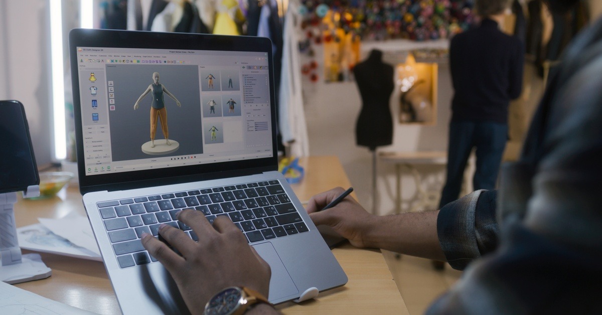 A man using a laptop that's propped up on a stand. He's looking at some digitized clothing while writing something down.