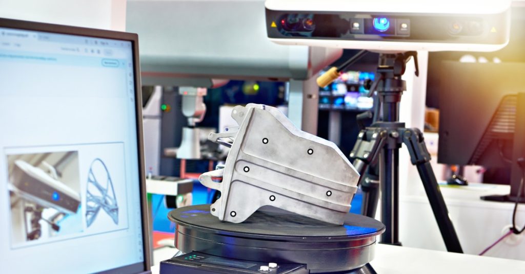 A metal component sitting on a pedestal while getting scanned by a laser scanner. There is a computer screen nearby.